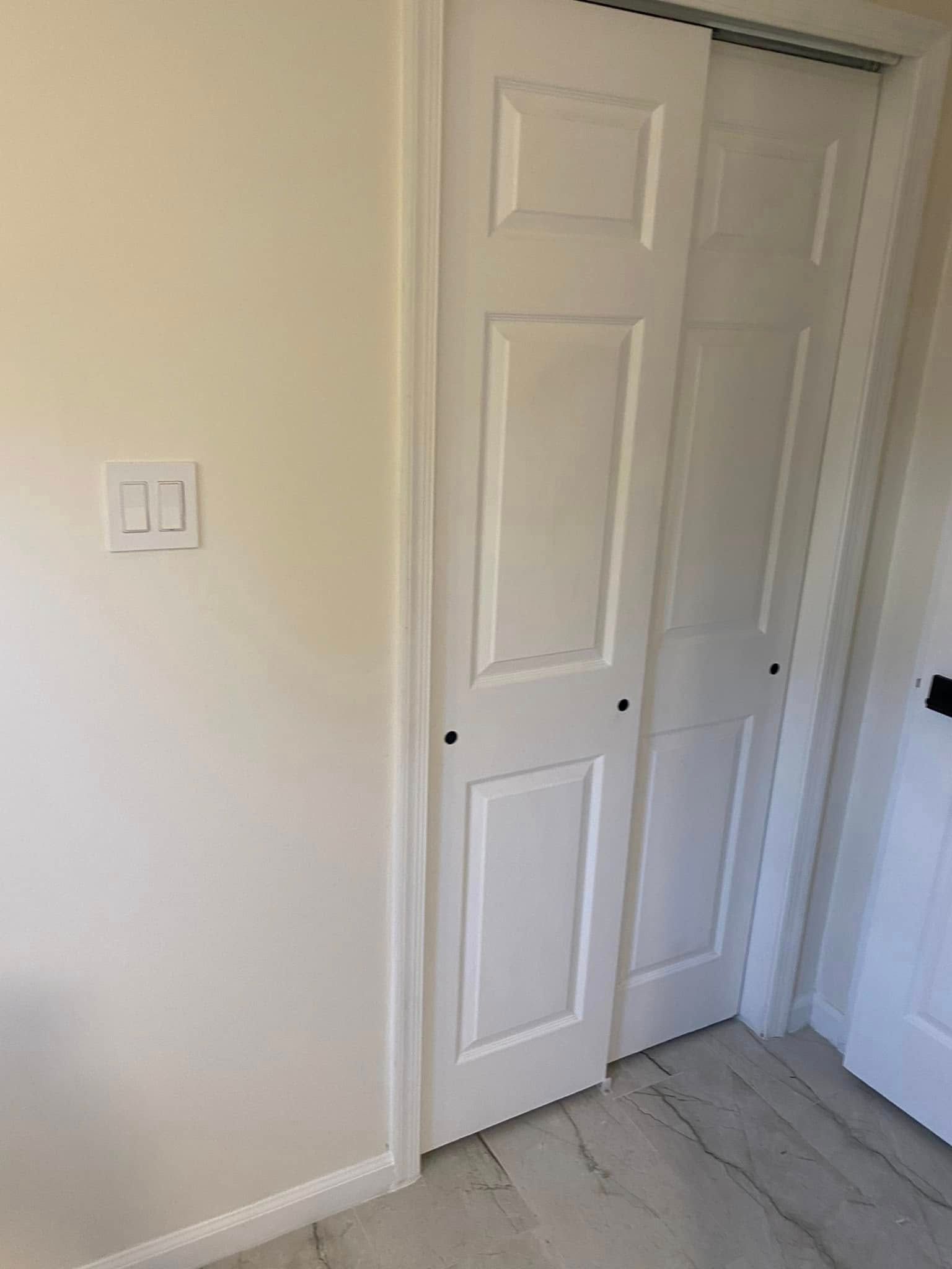 White bi-fold closet doors in a room with light-colored walls and flooring, a light switch is visible on the wall.