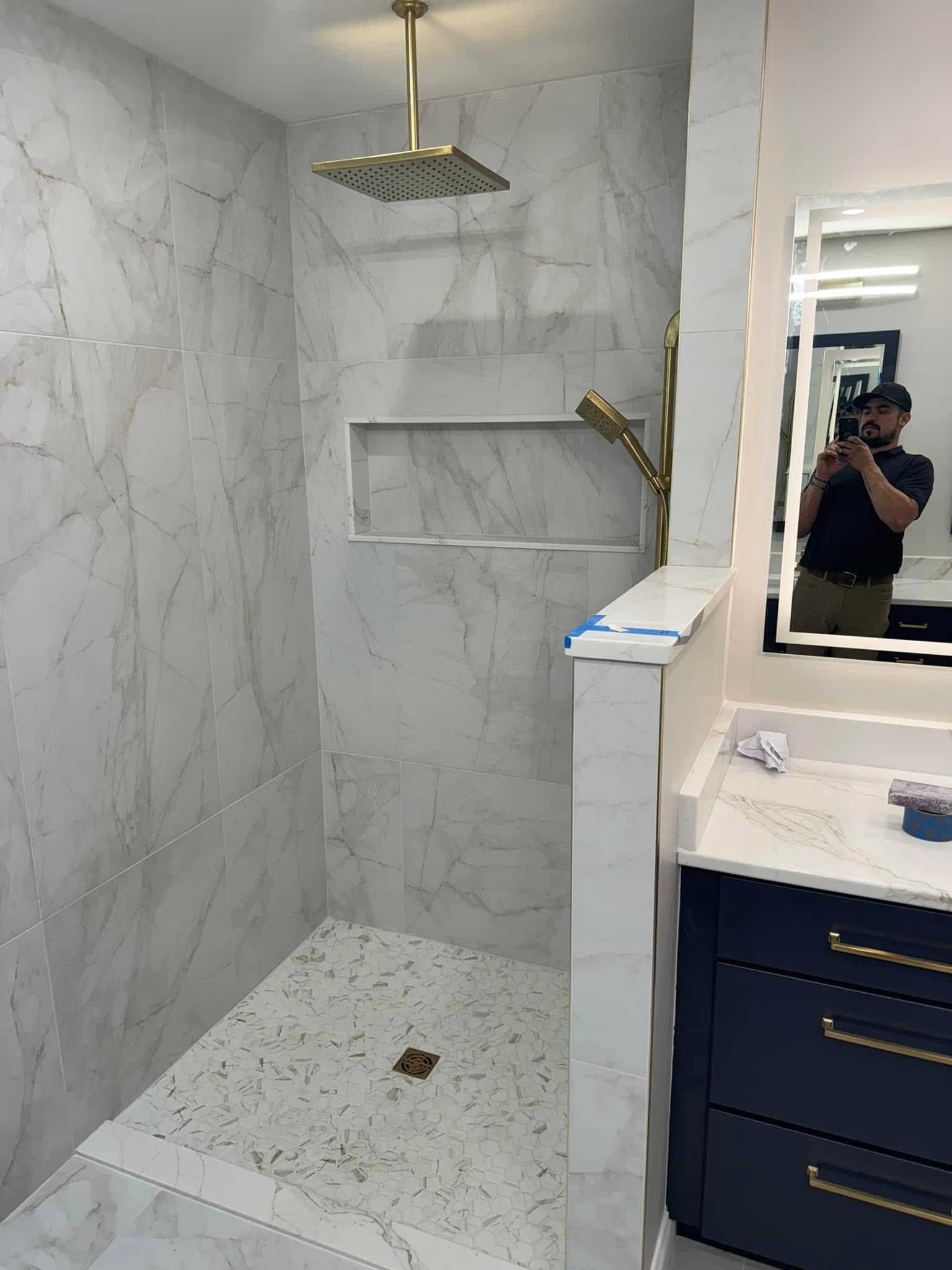 A newly tiled shower with gold fixtures and a recessed shelf. A person is reflected in the mirror taking the picture.