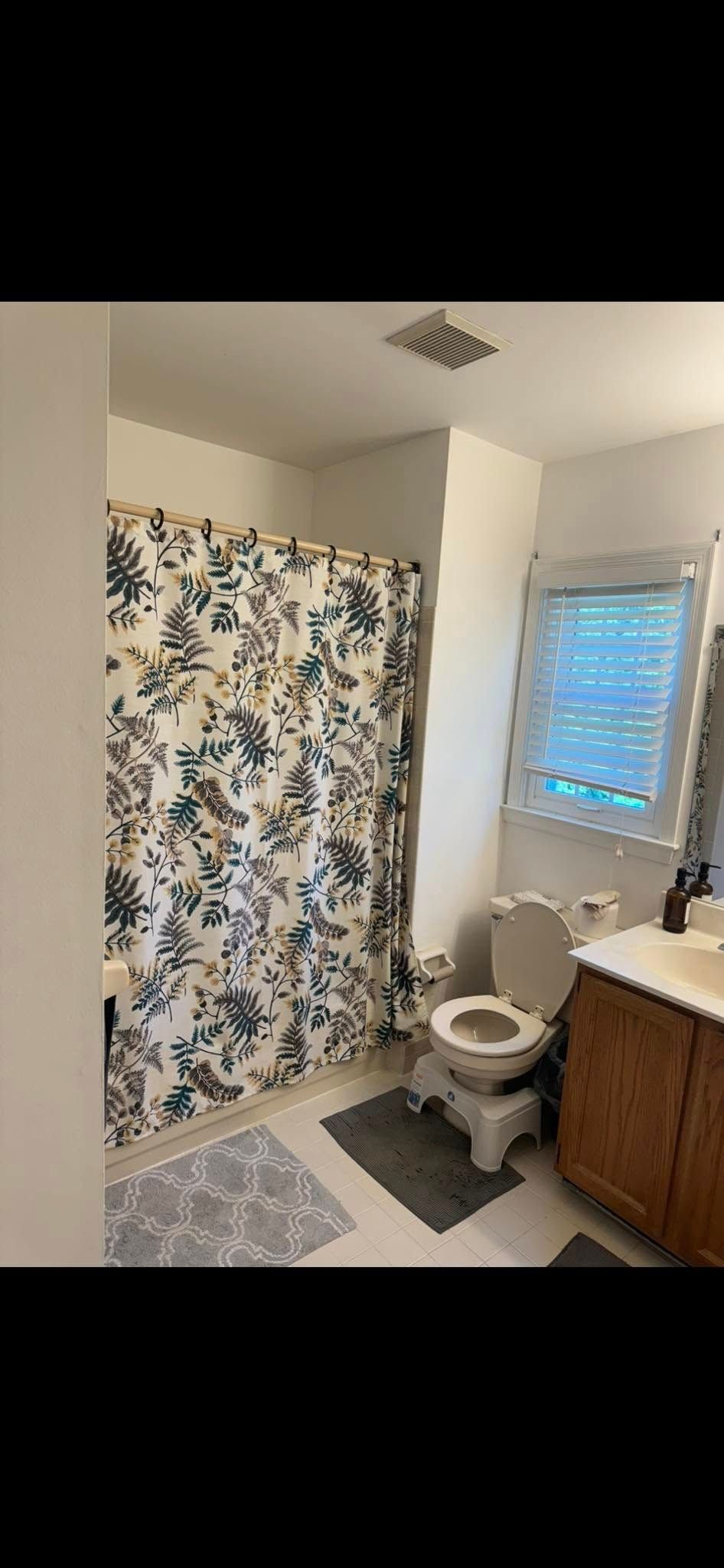 Bathroom with floral shower curtain, toilet with step stool, vanity, and window with blinds.