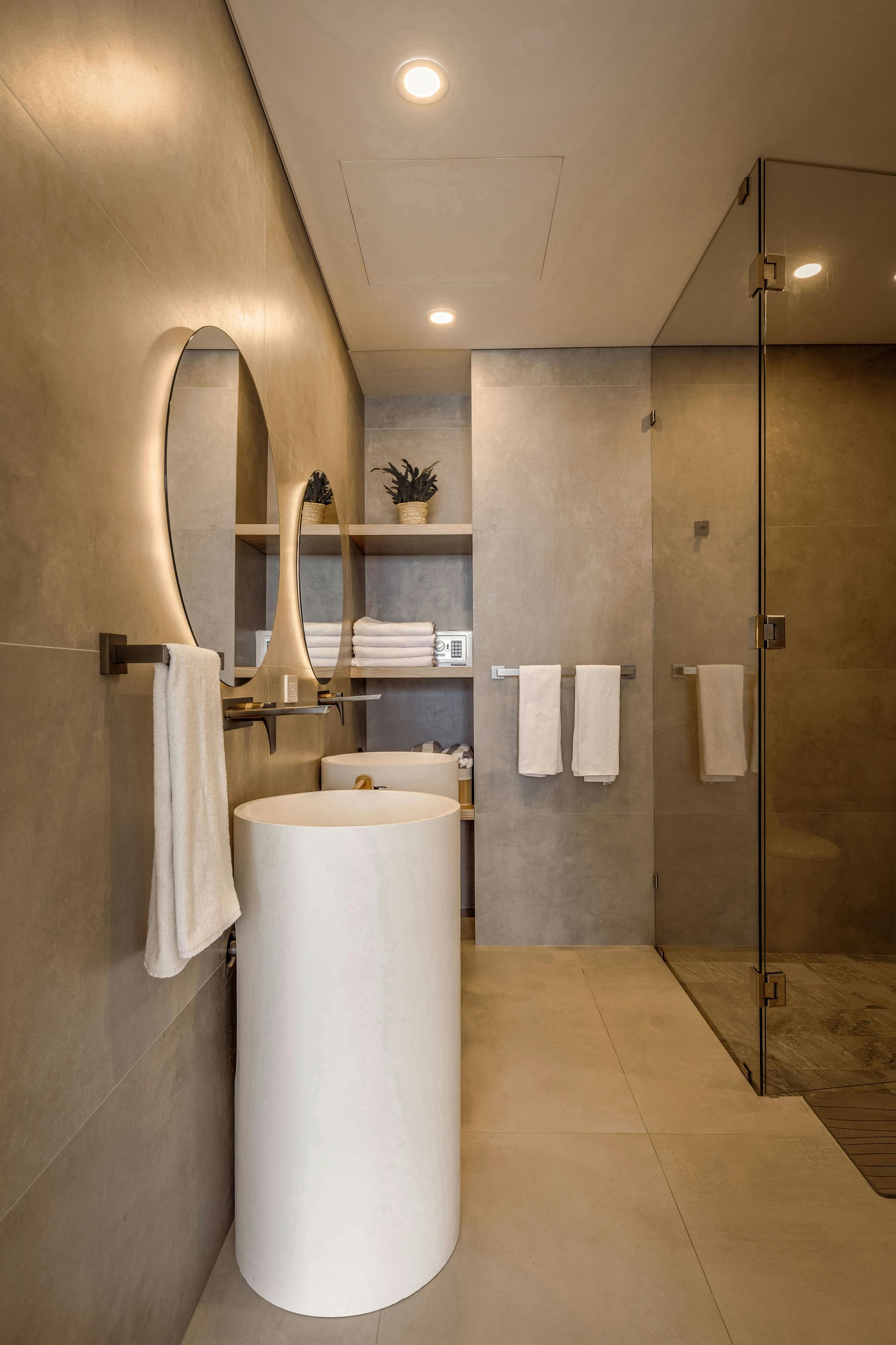 A modern bathroom with a cylindrical white pedestal sink, a circular backlit mirror, and a glass-enclosed shower stall.