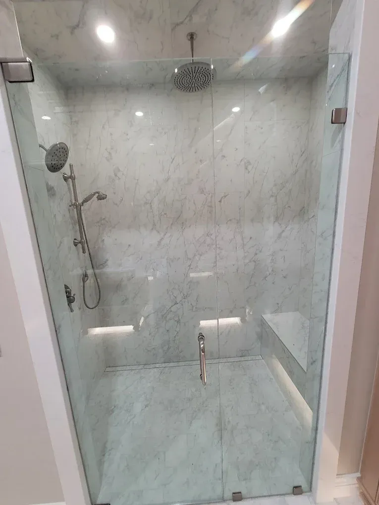 Glass-enclosed shower with marble walls, two shower heads, bench, and double doors.