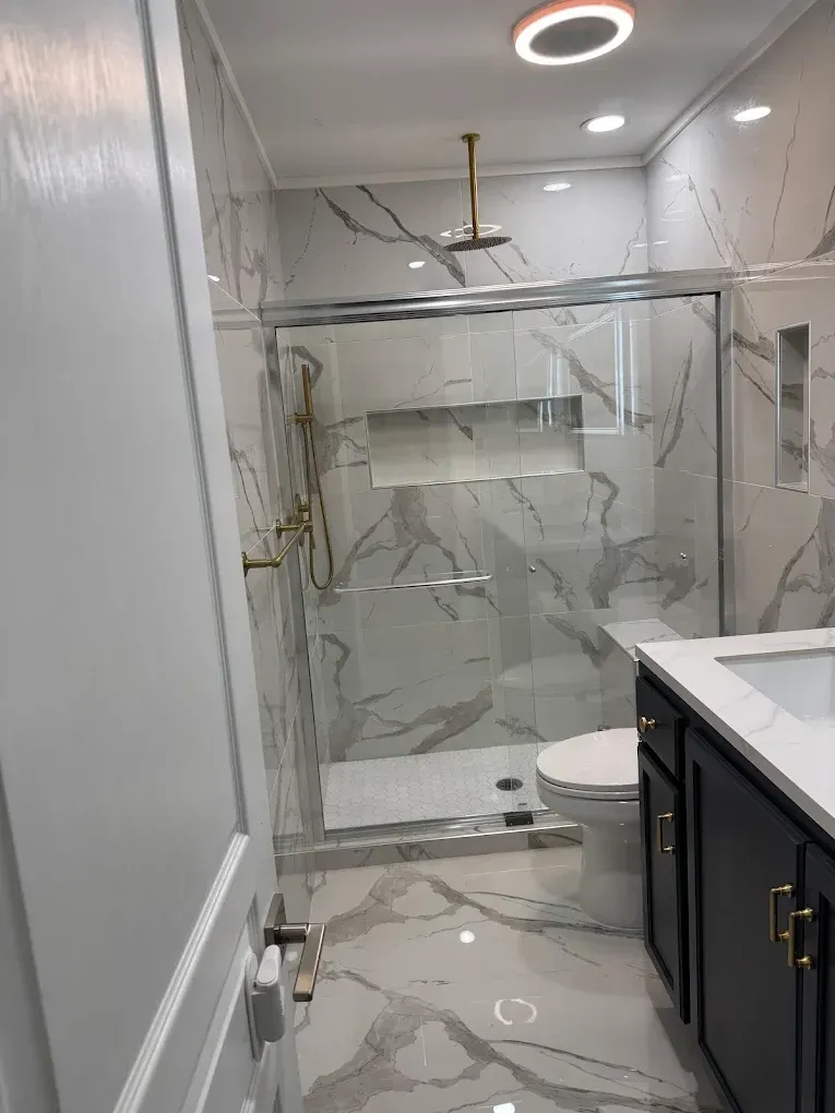 Newly renovated bathroom with marble-look tile, glass shower, gold fixtures, and dark vanity.