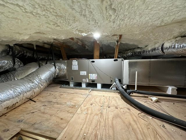 HVAC unit in an attic with ductwork and spray foam insulation. Wooden platform and rafters are visible.