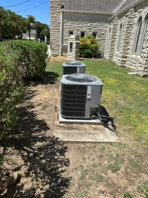 Two gray air conditioning units on concrete pads beside a stone building and overgrown shrubs.