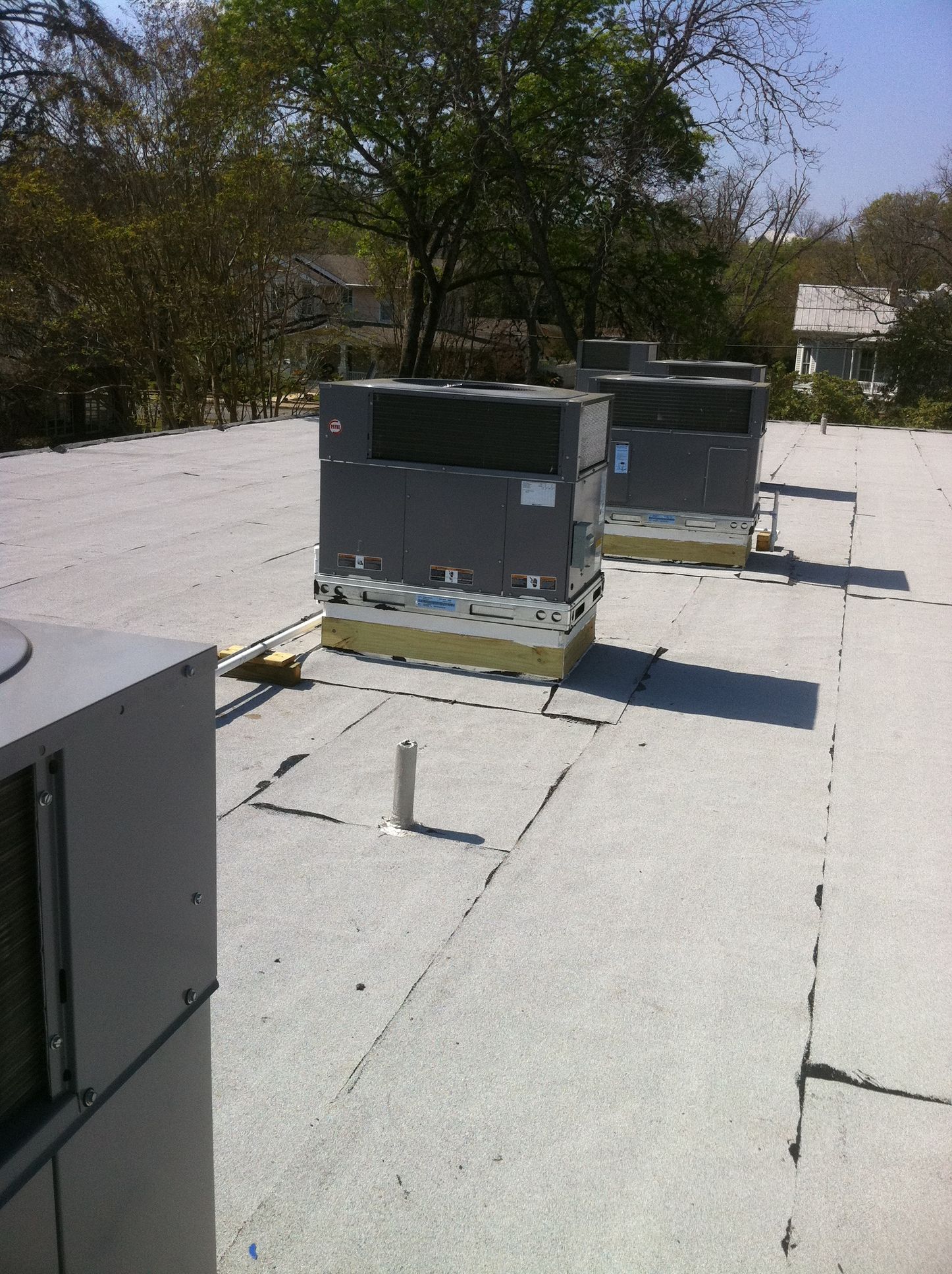 Rooftop HVAC units on a white flat roof, trees and houses in the background on a sunny day.