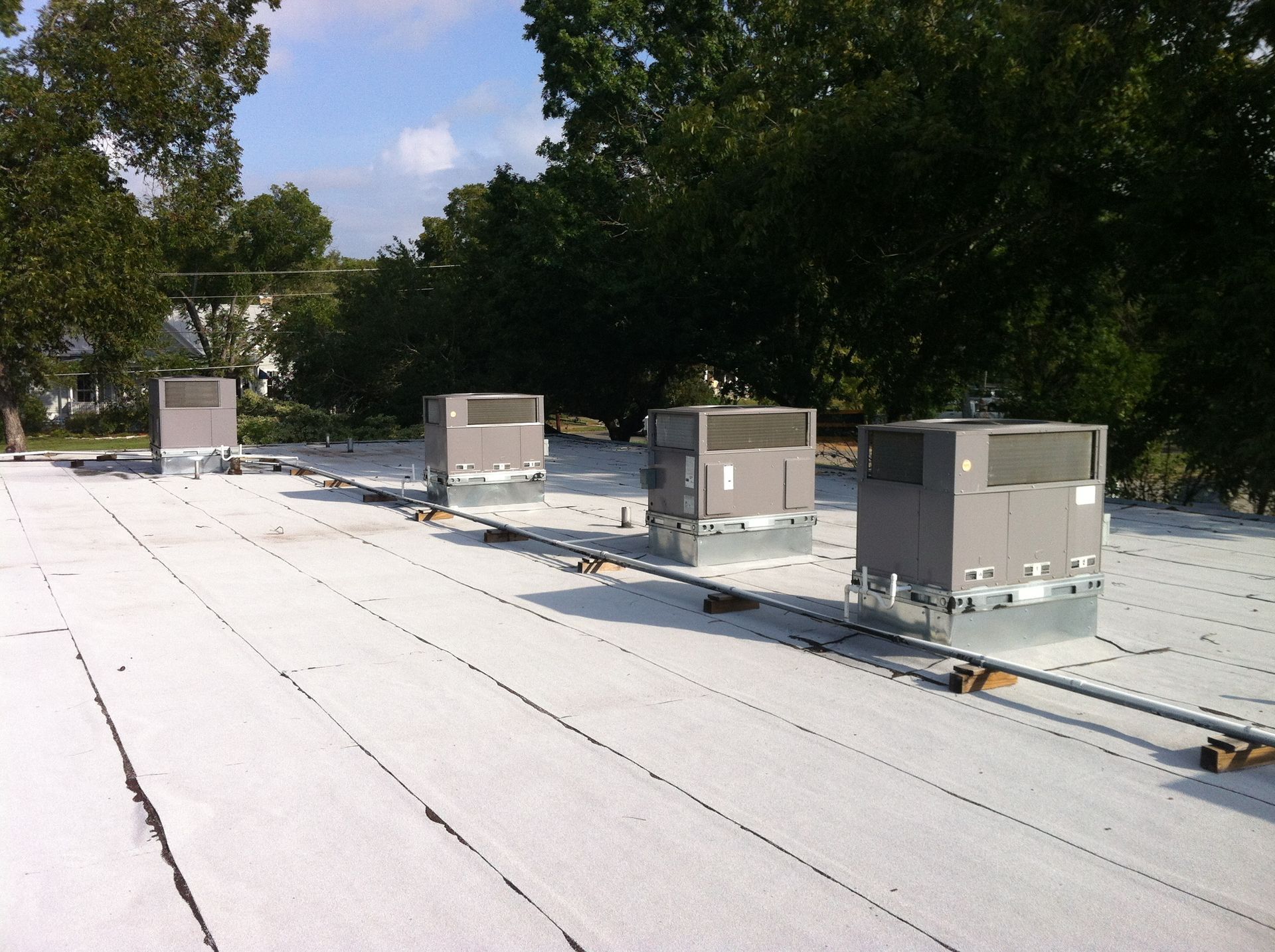 Four rooftop HVAC units on a white flat roof, with connecting pipes and trees in the background.