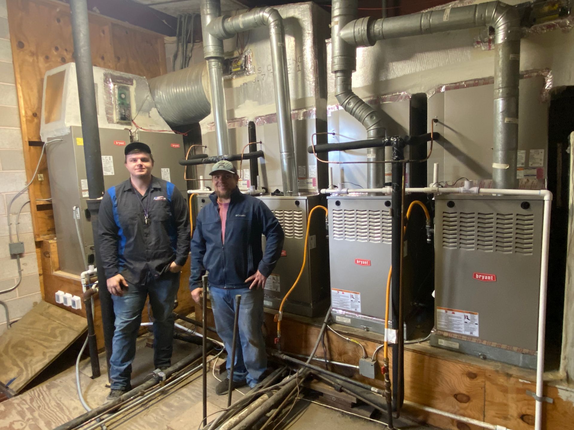 Two people stand beside new HVAC units in a utility room.