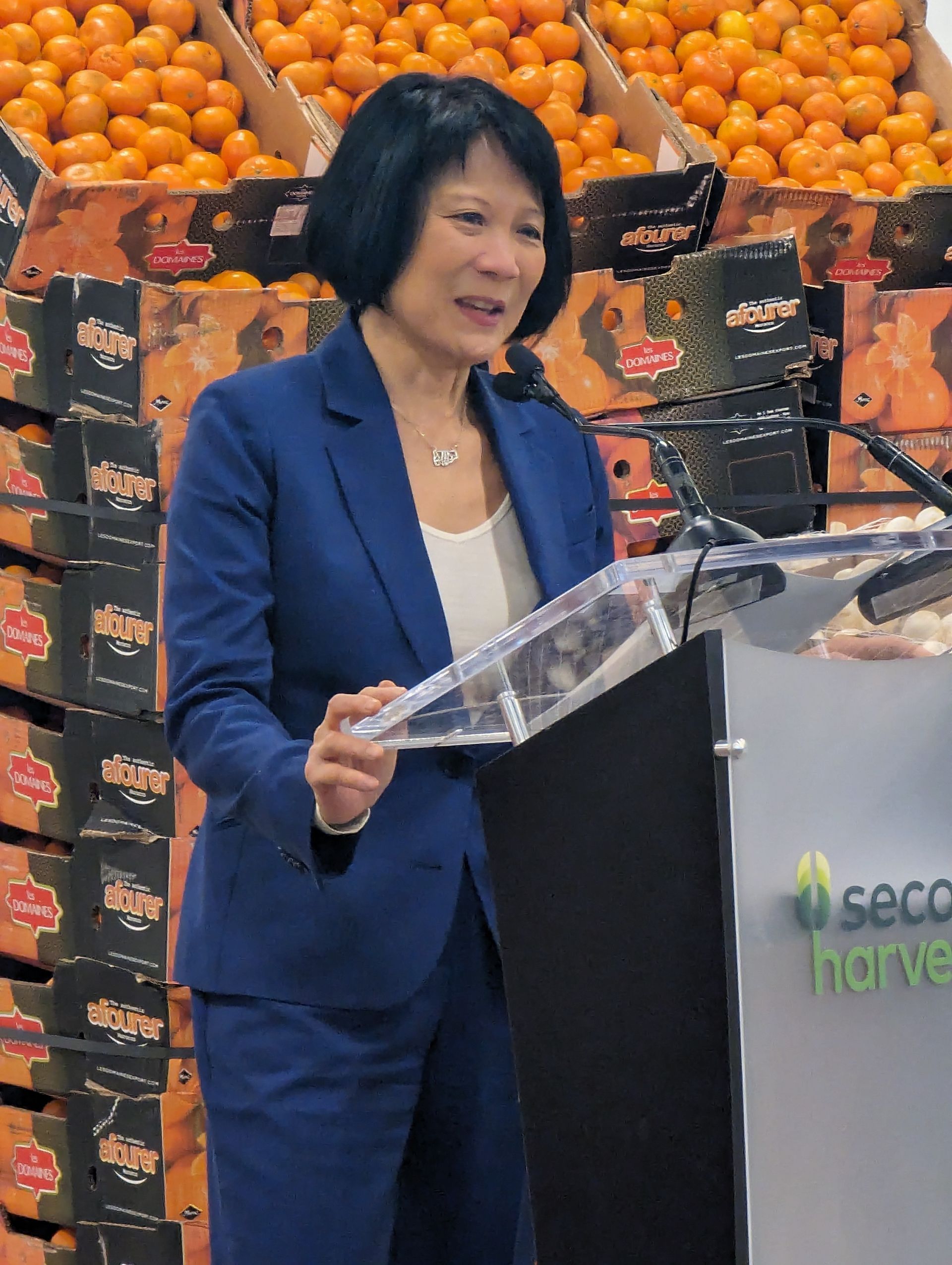 A woman in a blue suit is standing at a podium in front of boxes of oranges.
