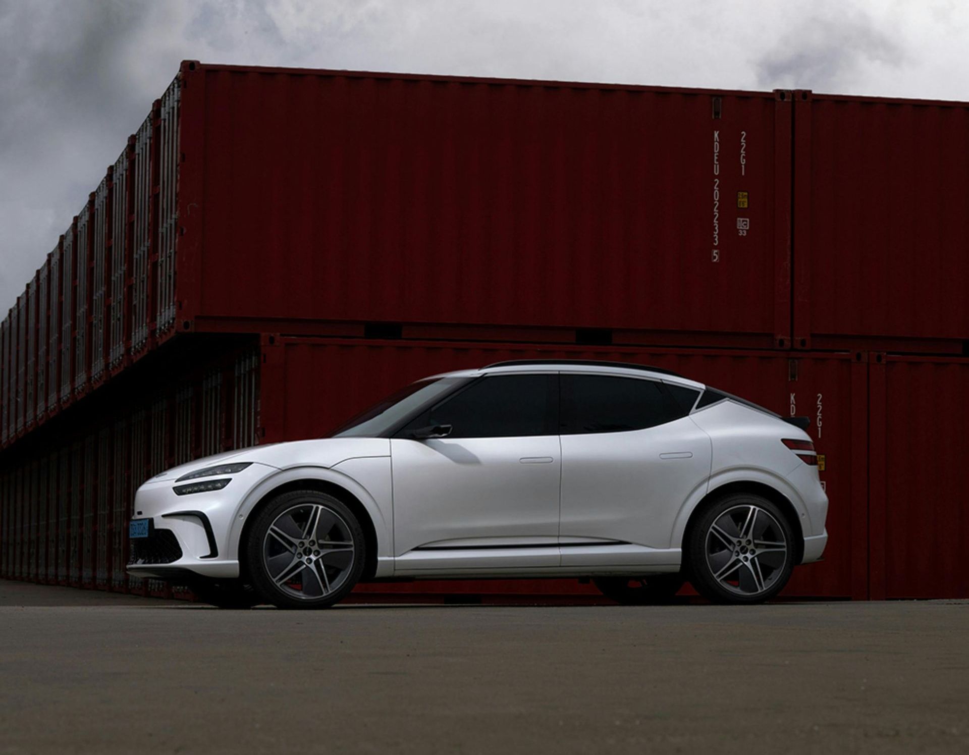 A white car is parked in front of a stack of red shipping containers.