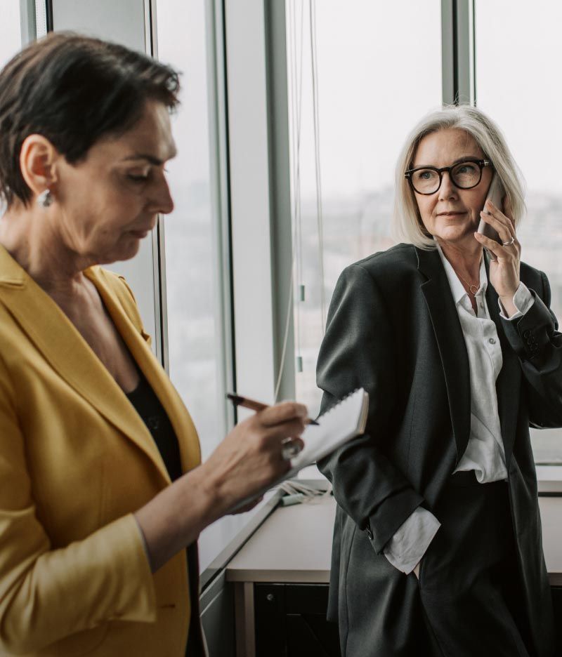 A woman in a yellow jacket is writing on a piece of paper while another woman talks on a cell phone.
