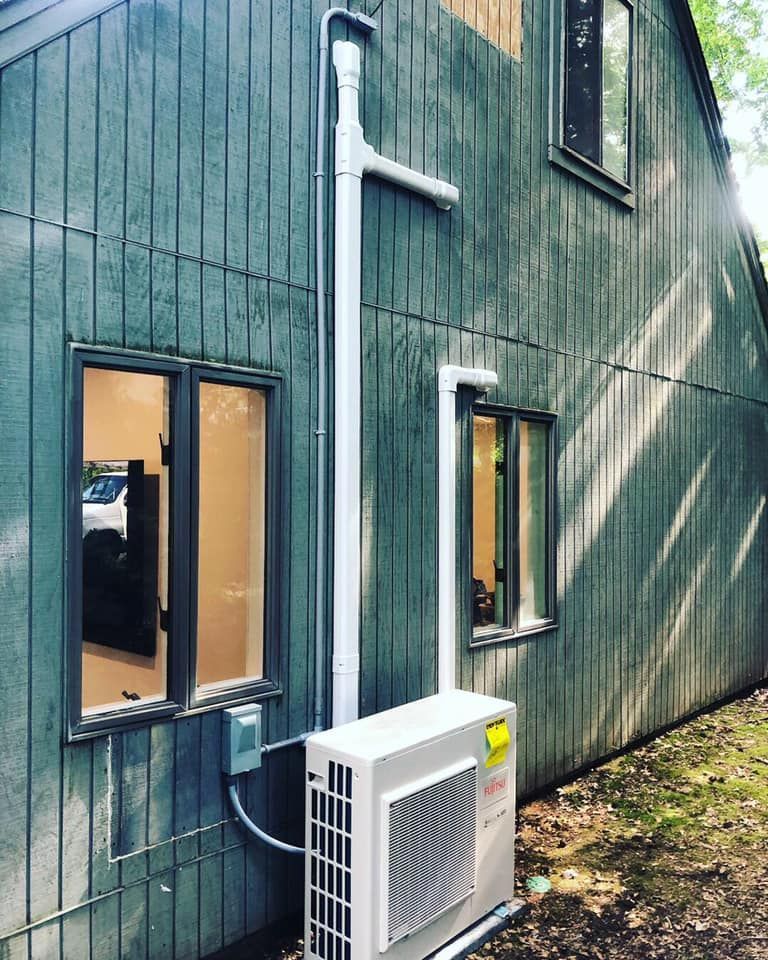 An air conditioning unit mounted on a gray-green house exterior, with white ductwork and two windows.