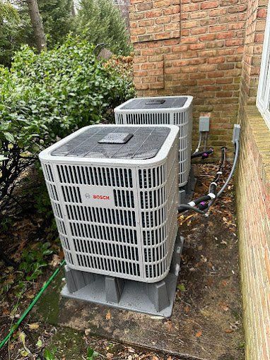 Two Bosch air conditioning units against a brick wall, on concrete bases, next to green foliage.
