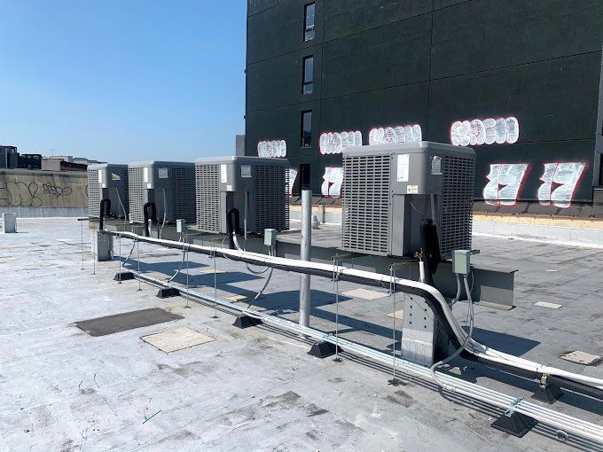 Rooftop view of several air conditioning units with electrical conduit and a building under a blue sky.