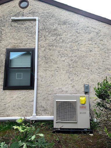 An outdoor HVAC unit on a stucco building with a window and white pipes.