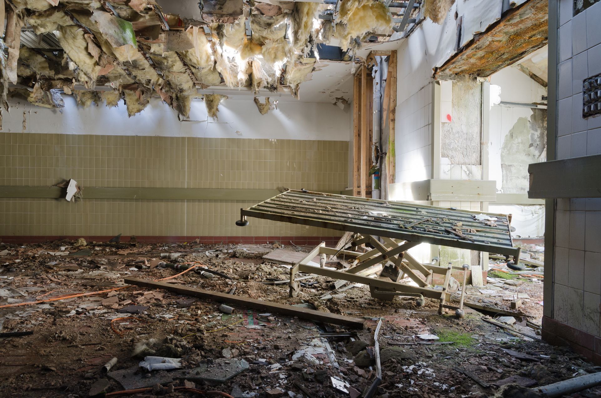 Interior of a decaying room with collapsed ceiling and debris on the floor, medical table in the center.