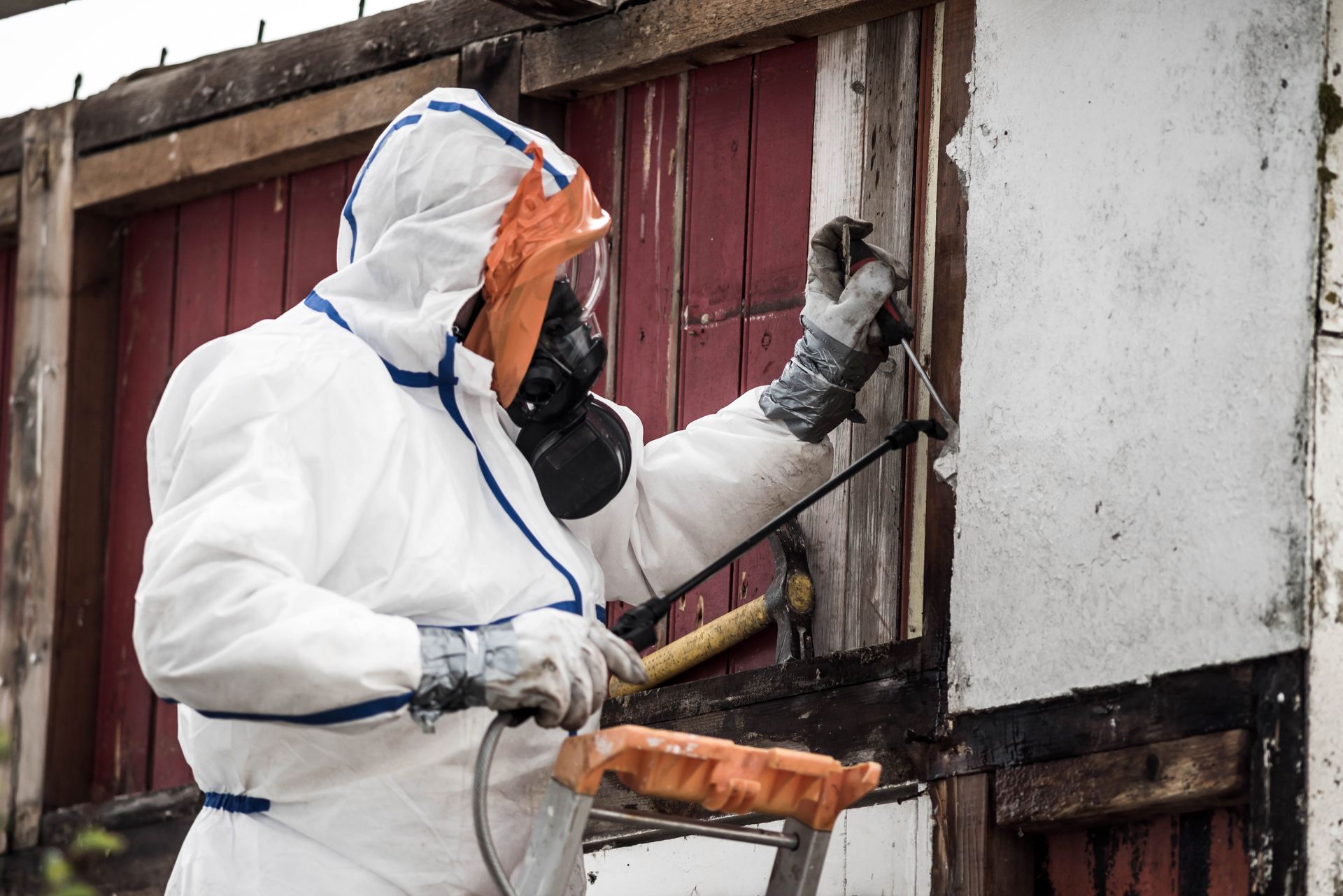 Person in protective suit spraying a building, wearing a respirator, outdoors.