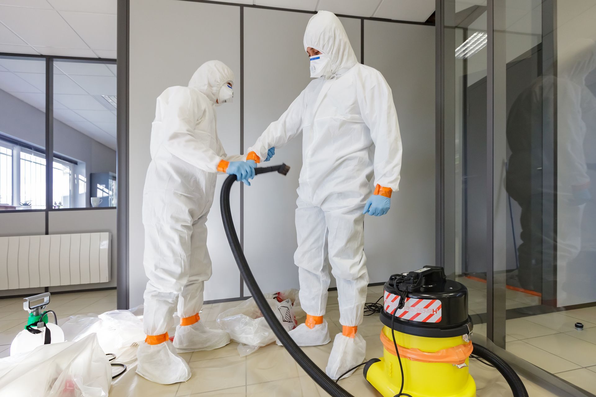 Two people in white hazmat suits using a vacuum cleaner in an office.