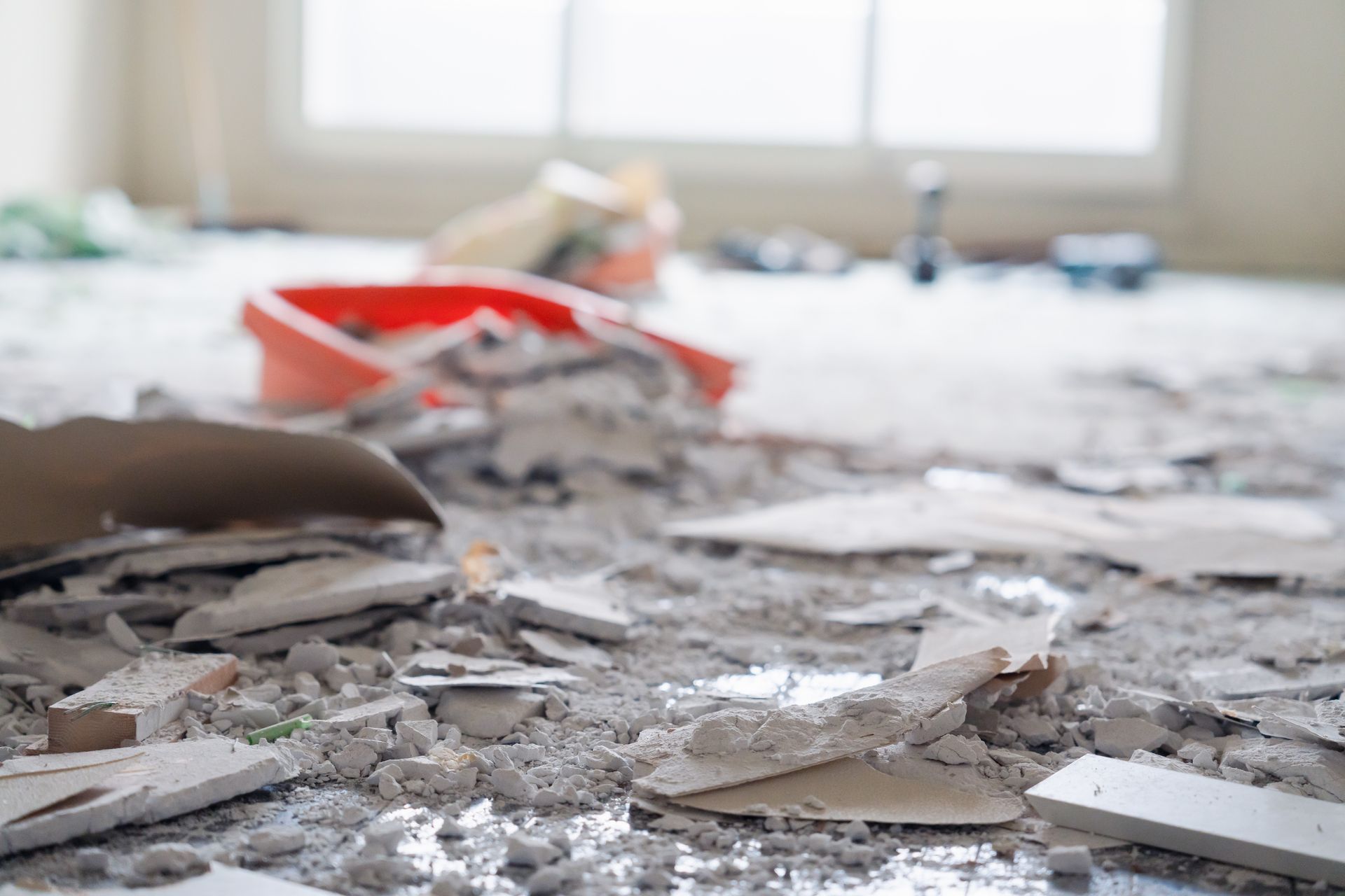 Demolition debris covers a room floor with a red bucket in the background by a window.