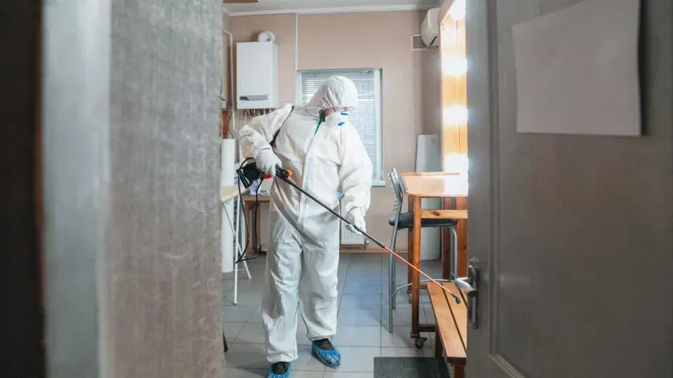 Person in white protective suit kneels near a wall, bucket, and tool.