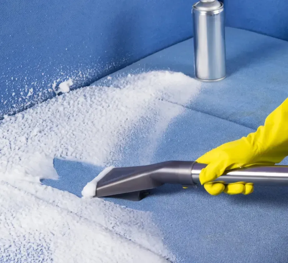 A person wearing yellow gloves is cleaning a blue couch with a vacuum cleaner.