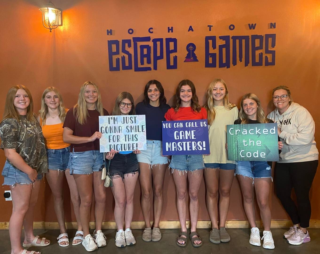 Group posing in front of Escape & Games sign, holding coding-themed team signs.
