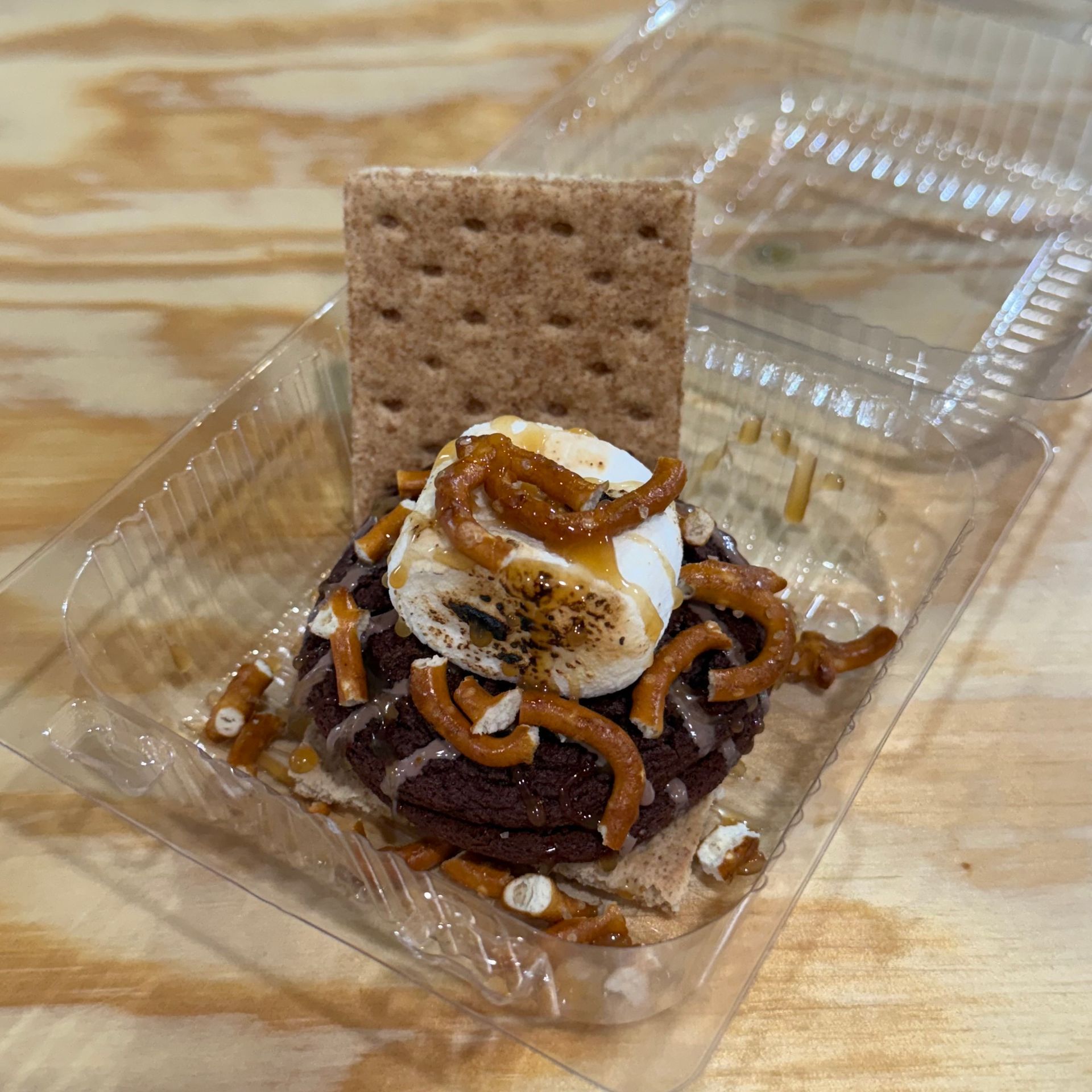 Takeout dessert with whipped cream, caramel drizzle, pretzels, and a graham cracker in a plastic container