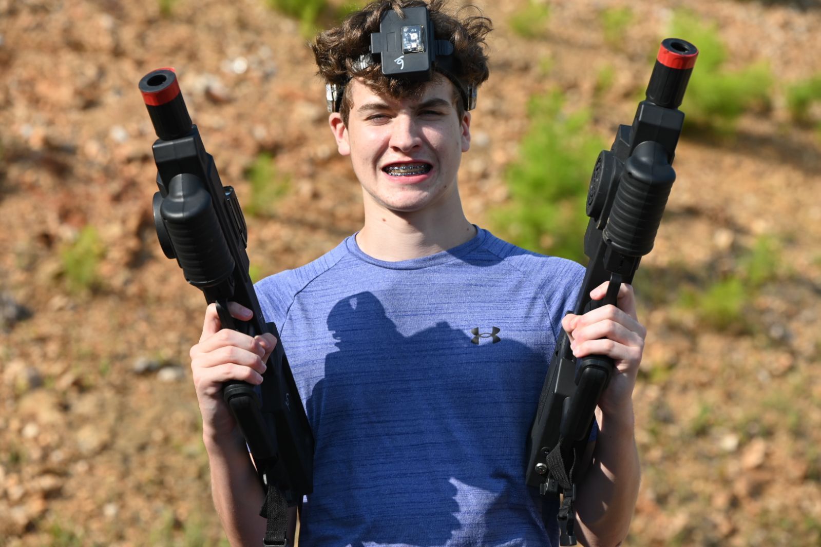 Teen holding two toy blasters outdoors, wearing a head-mounted camera and smiling.