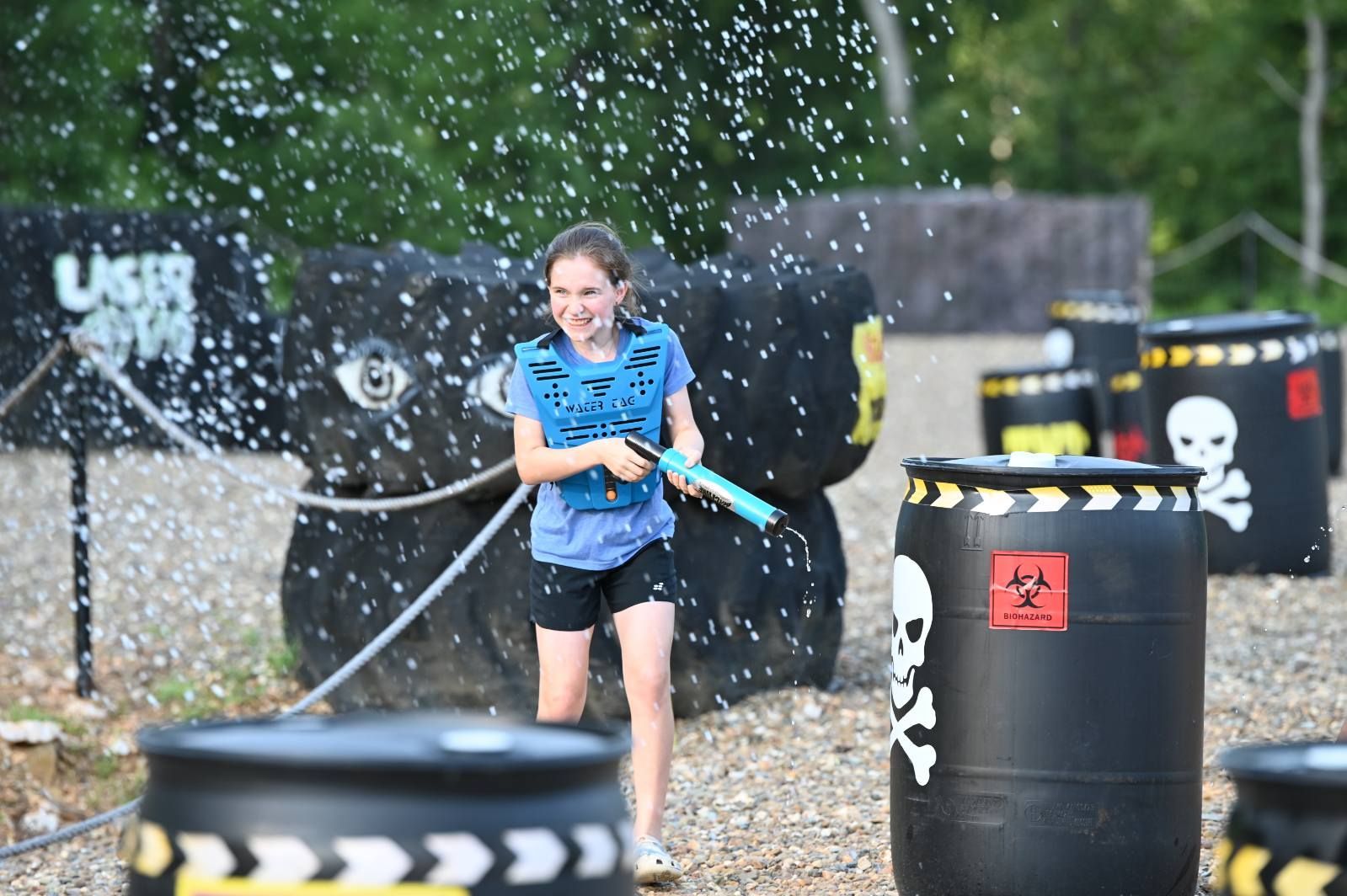 Child in blue shirt smiling while holding a water gun in a splashing outdoor paintball field