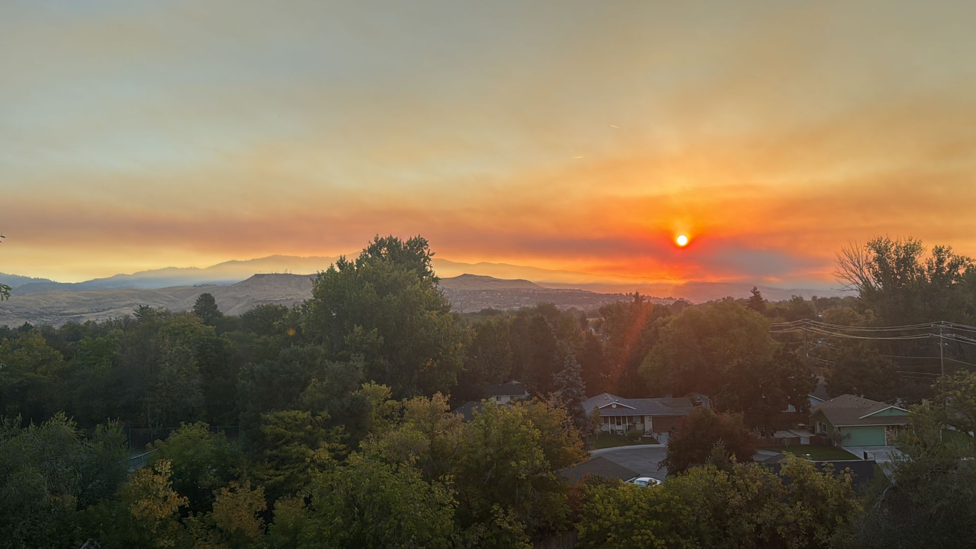 A sunset over a city with trees in the foreground and mountains in the background.