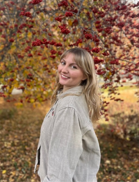 Woman with glasses smiles, leaning against a tree, arms crossed, wearing a light sweater.