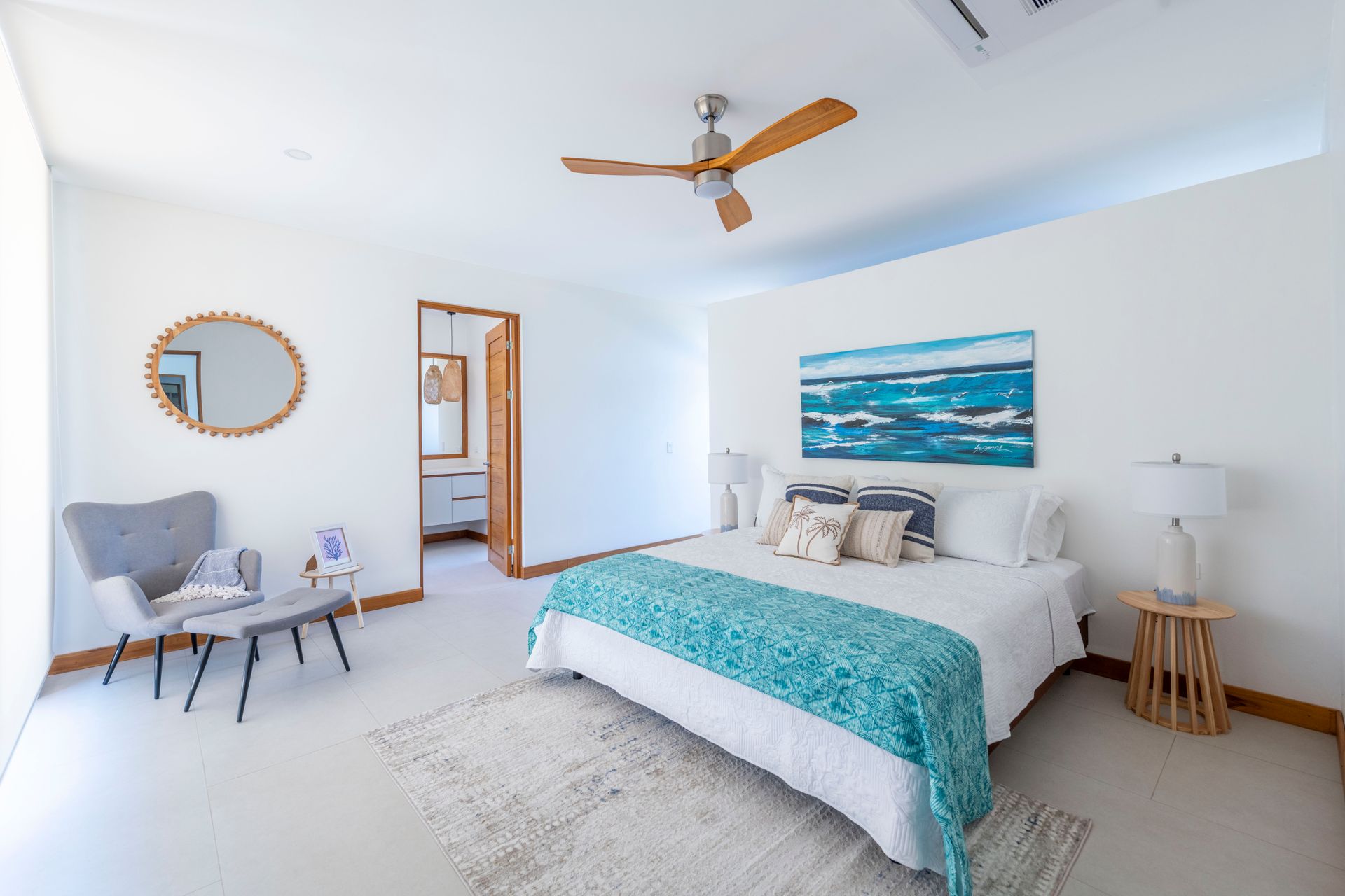 Bright bedroom with white walls, bed with teal throw, armchair, and view into bathroom.