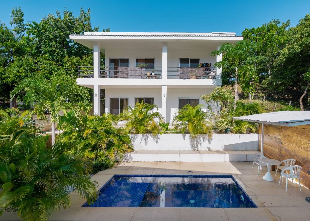White two-story house with balconies, a pool, and lush tropical vegetation under a blue sky.