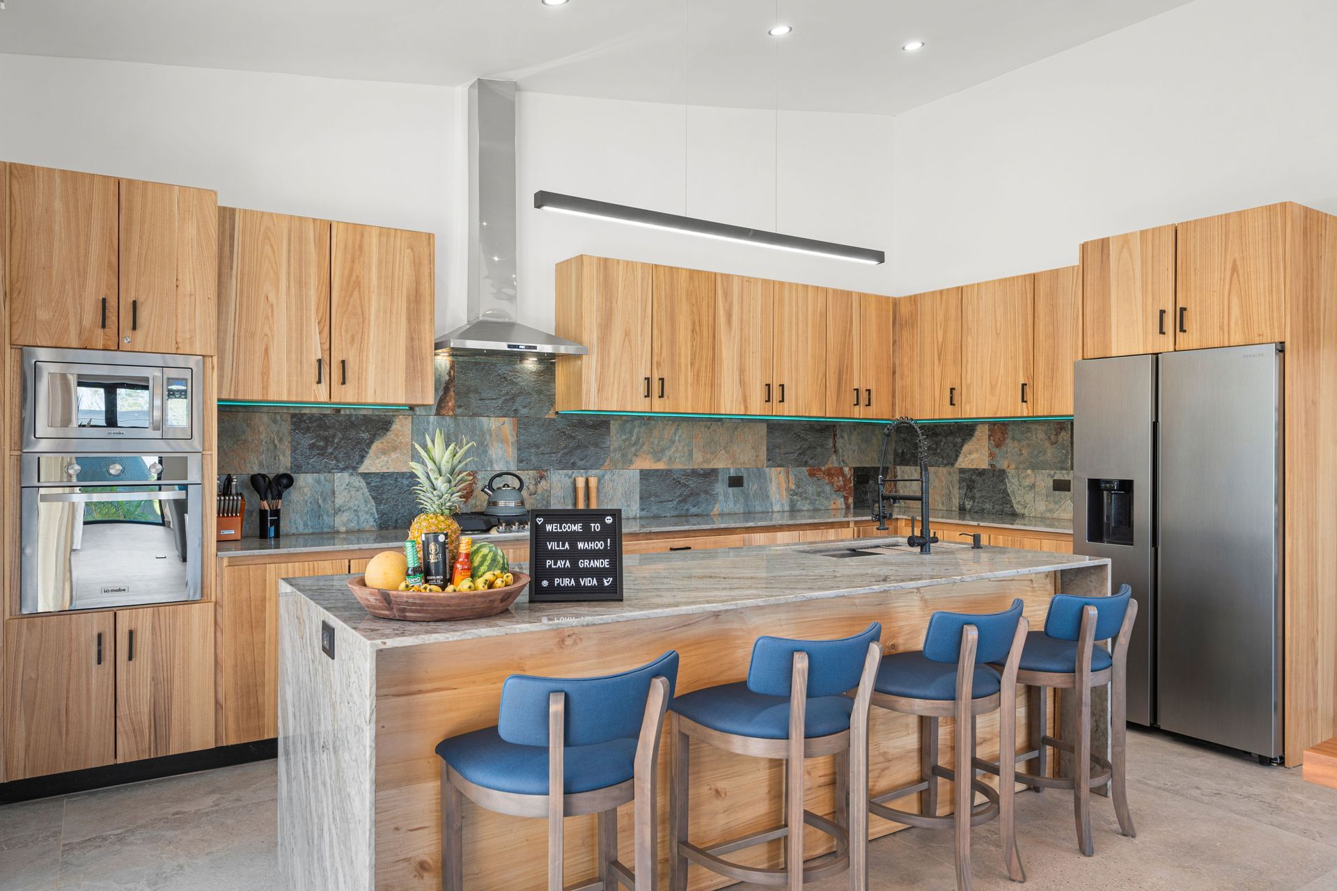 Modern kitchen with wooden cabinets, stone backsplash, island with blue bar stools, and stainless steel appliances.