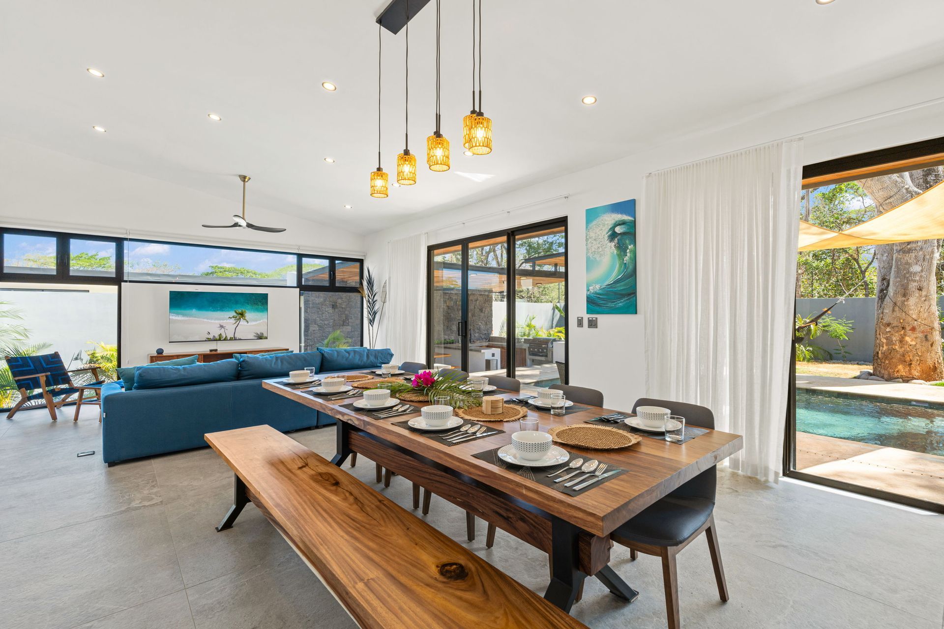 Open-concept dining and living area with long wooden table, blue sofa, and pool view.
