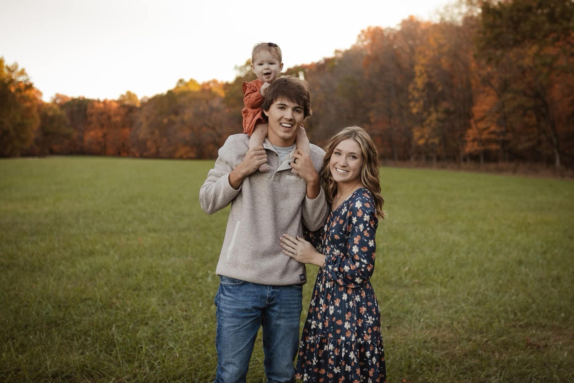 Family of three smiling in a field. Man holds child on shoulders. Woman in floral dress, autumn trees.