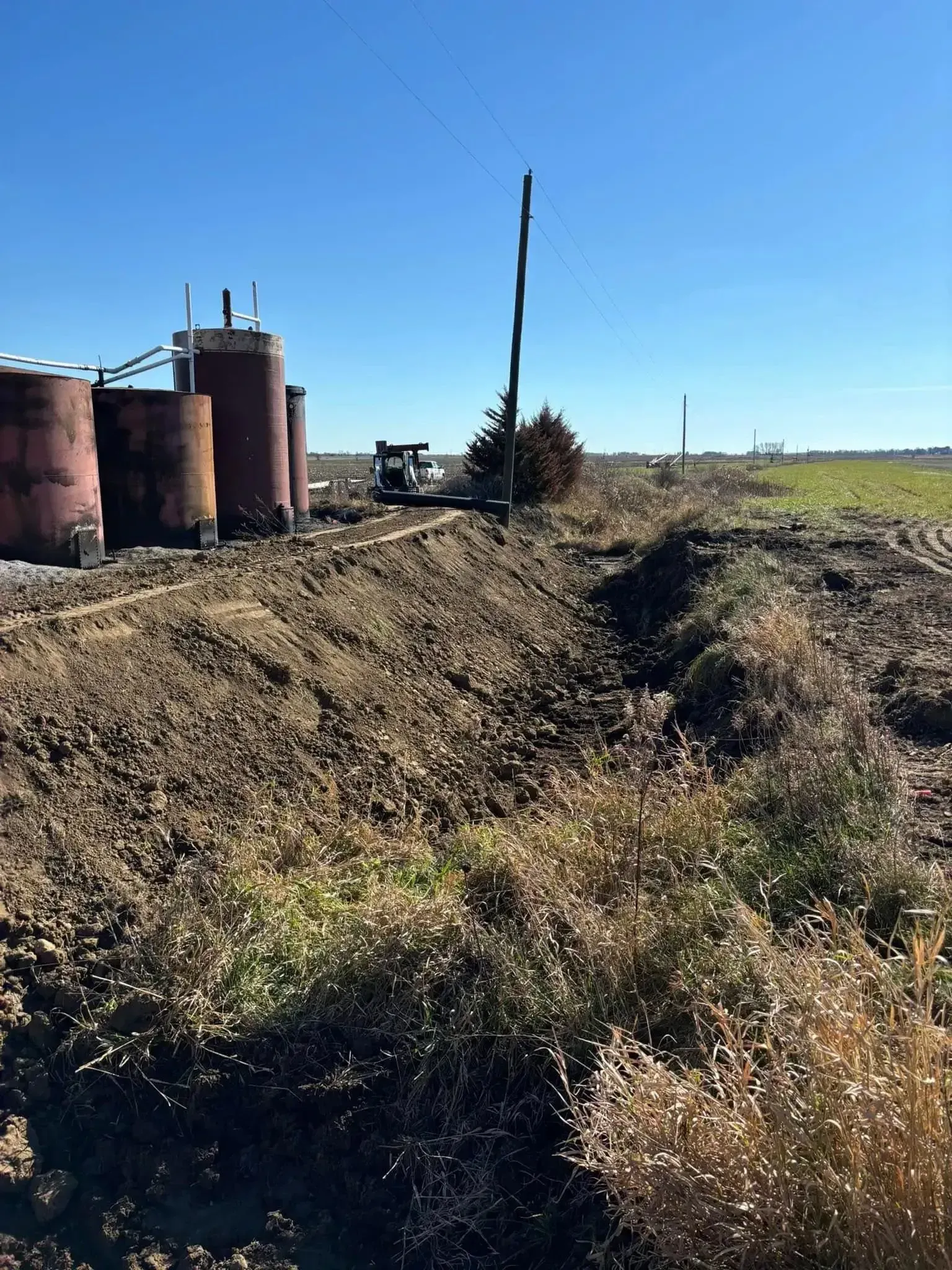 Brown tanks and oil pump near a dirt ditch and field under a blue sky.