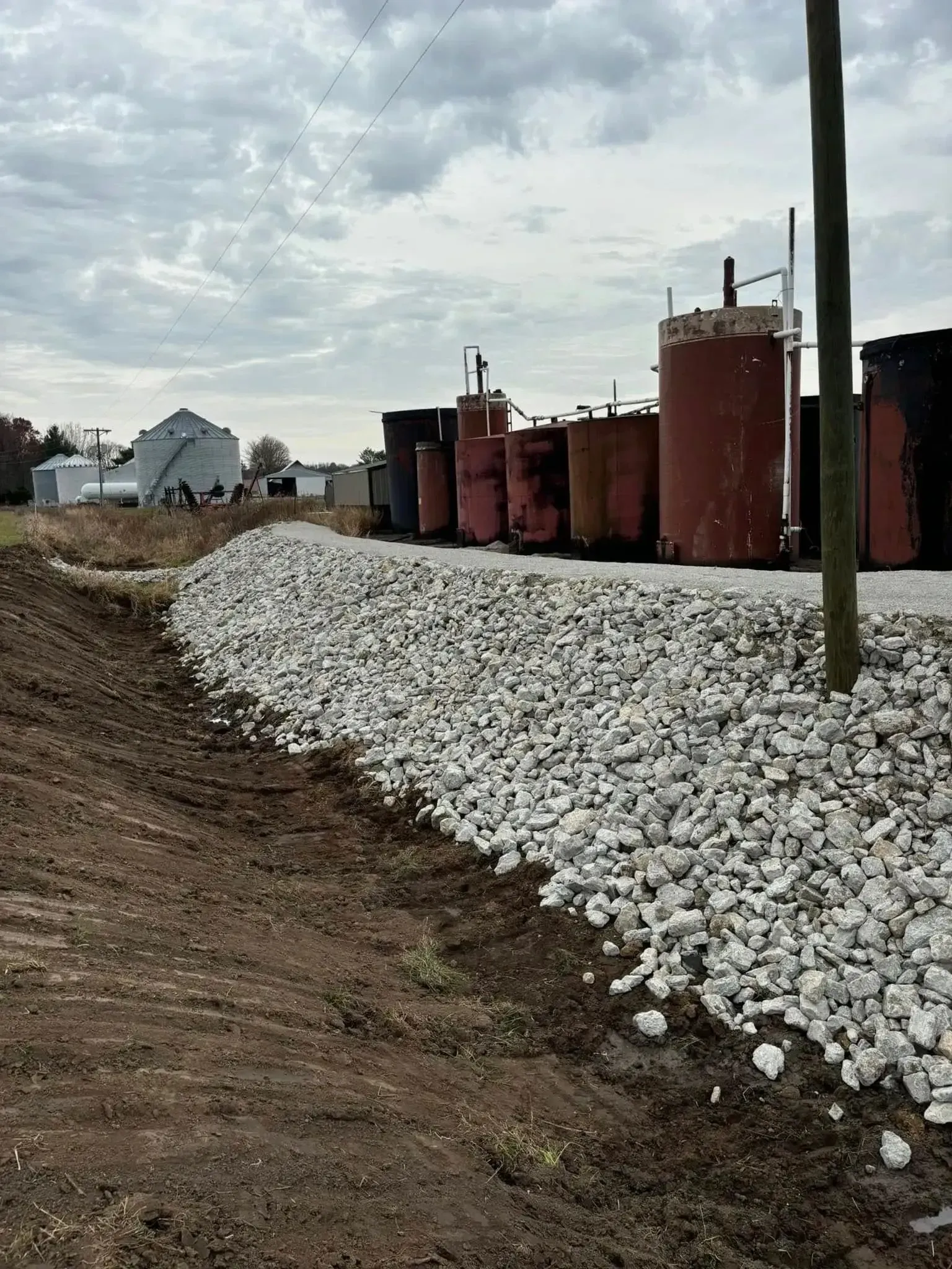Gravel-lined drainage ditch beside a row of storage tanks in a rural setting. Overcast sky.