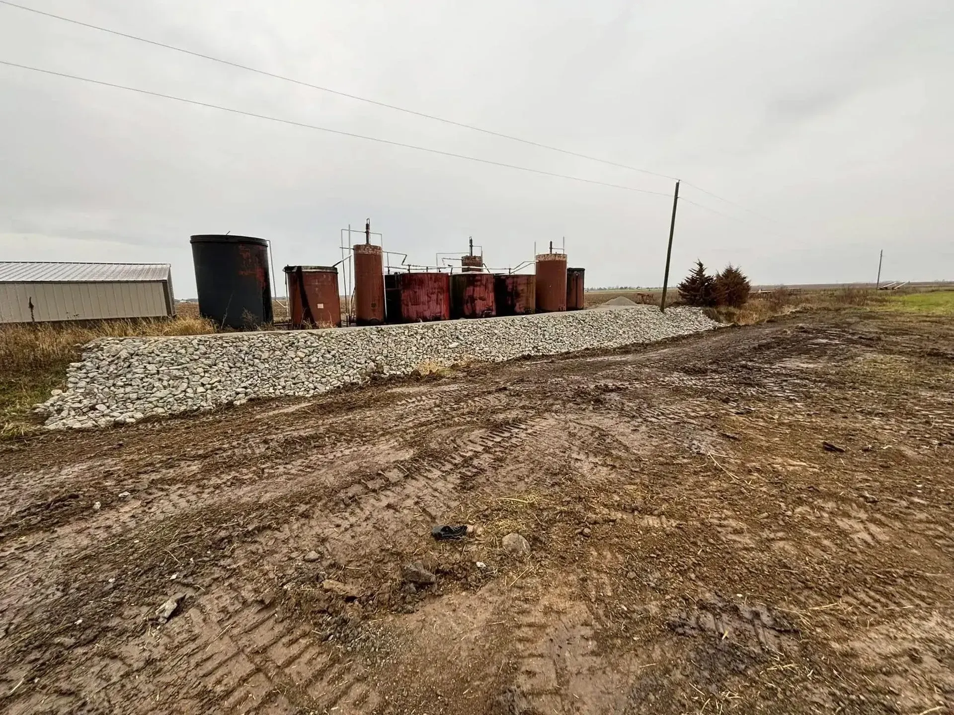 Old rusty tanks on a gravel berm in a field under a cloudy sky.