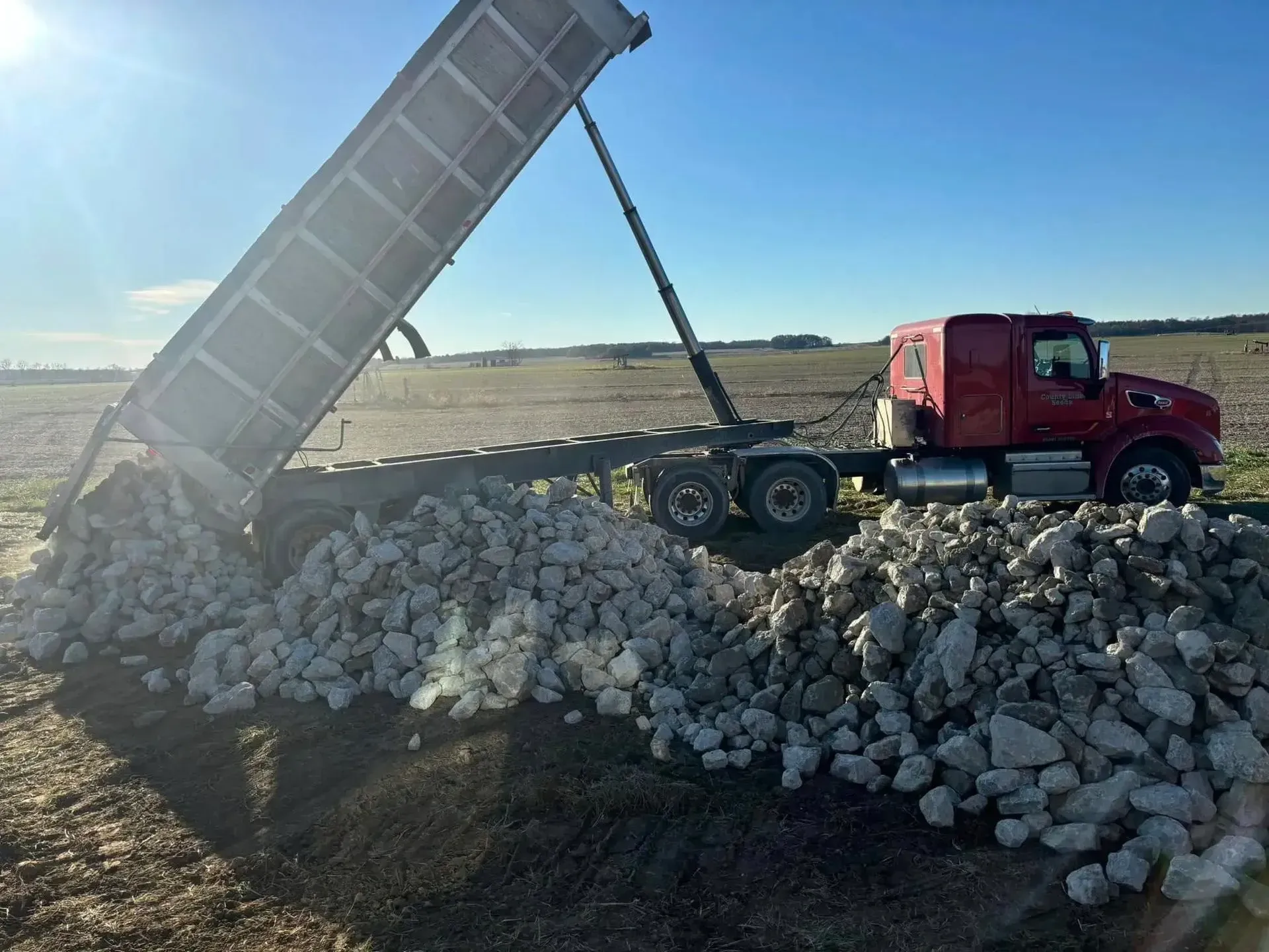 Red dump truck unloading rocks in a field on a sunny day.
