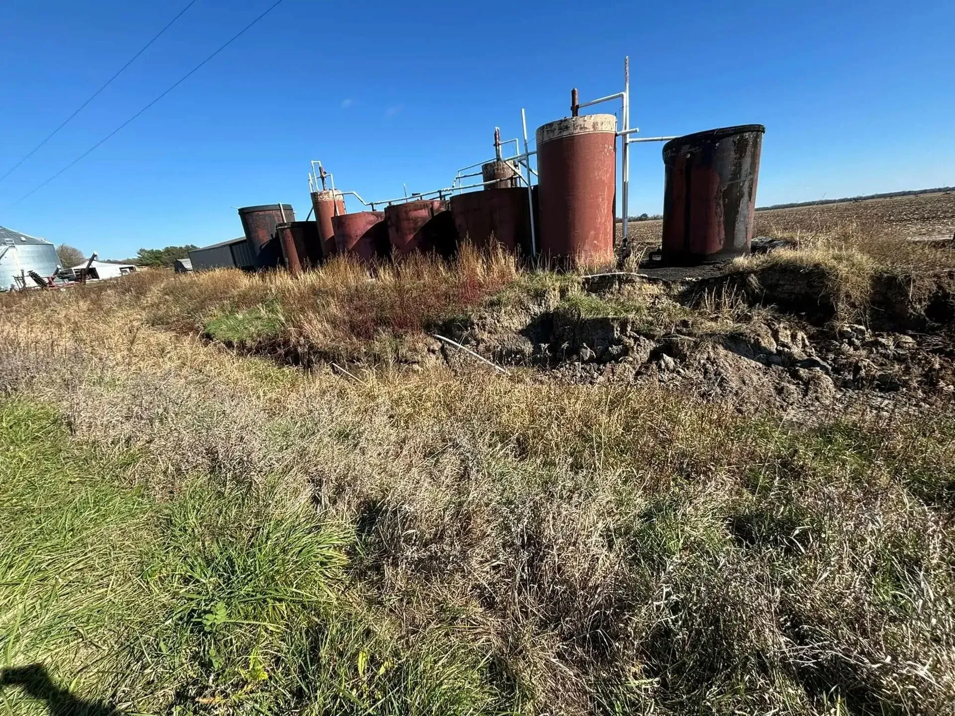 Old, rusty oil tanks on a grassy field against a clear blue sky.