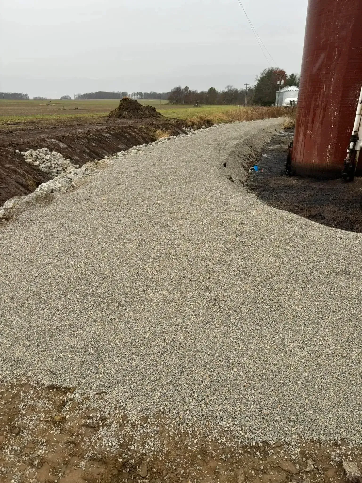 Gravel pathway curves around a red silo, leading towards a field under an overcast sky.