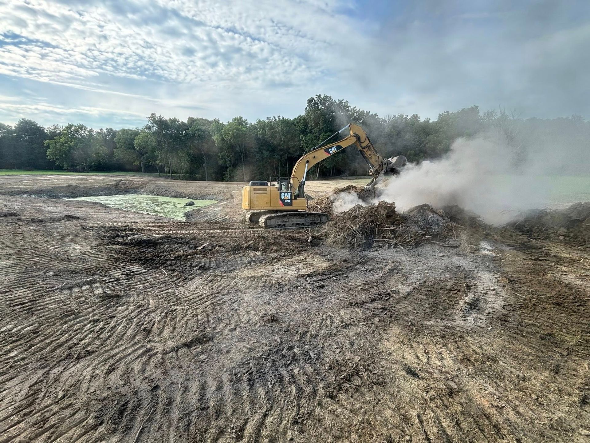 Yellow excavator moving debris, smoke rising, in an open field; trees in the background.
