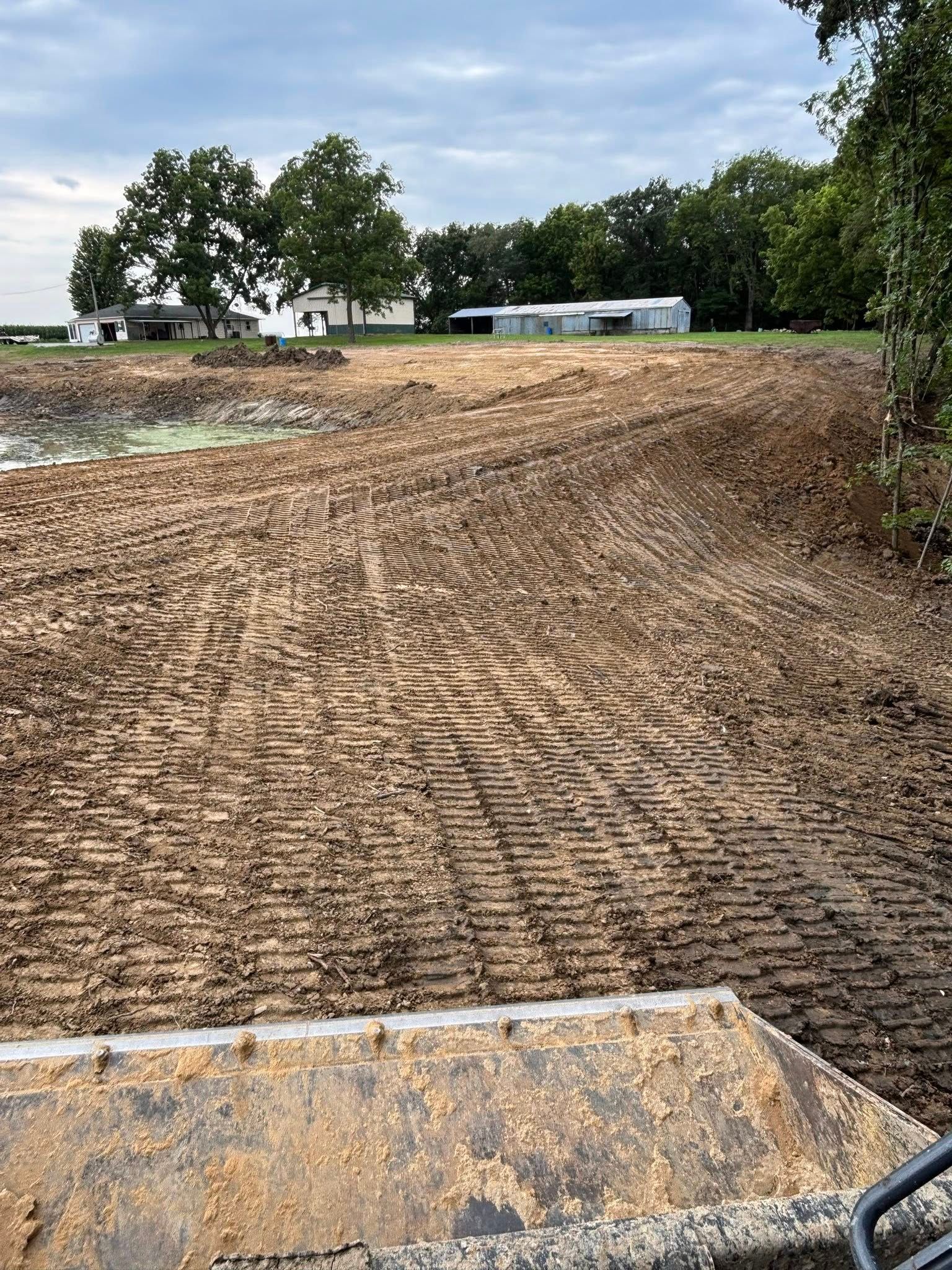 Dirt-covered ground near a body of water, with a building and trees in the background under a cloudy sky.