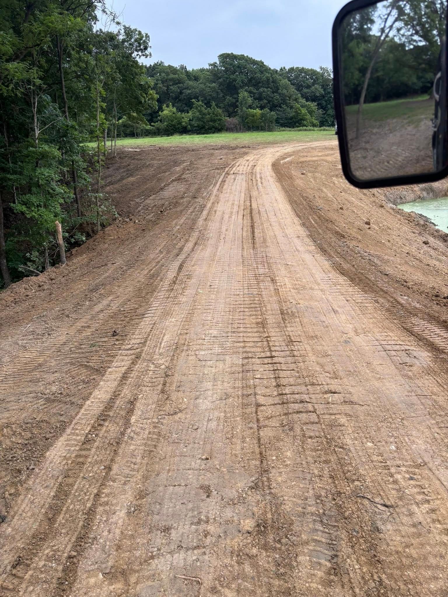 Dirt road leading into a green field, viewed from a vehicle. Side mirror in the frame.