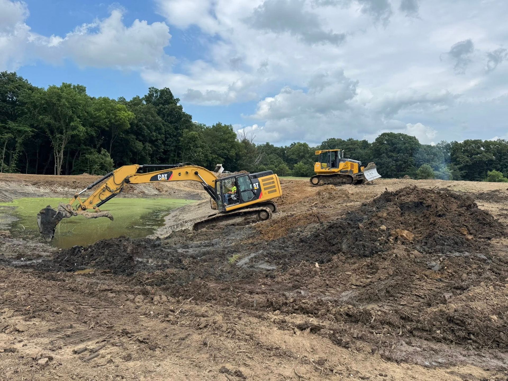 Construction site: Excavator and bulldozer moving earth under a cloudy sky. Green trees in the background.
