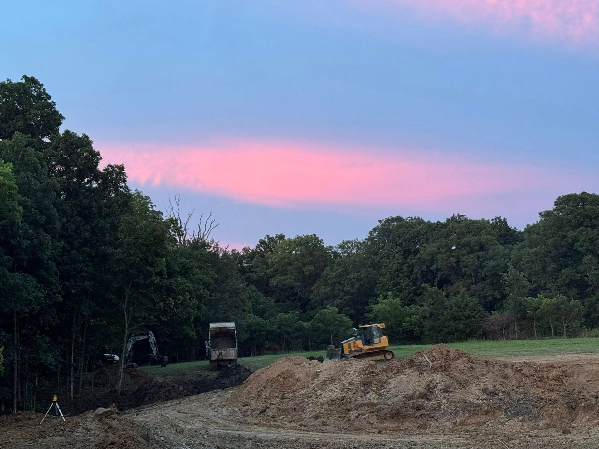 Construction site with earth-moving equipment under a pink and blue sunset sky; surrounded by trees.