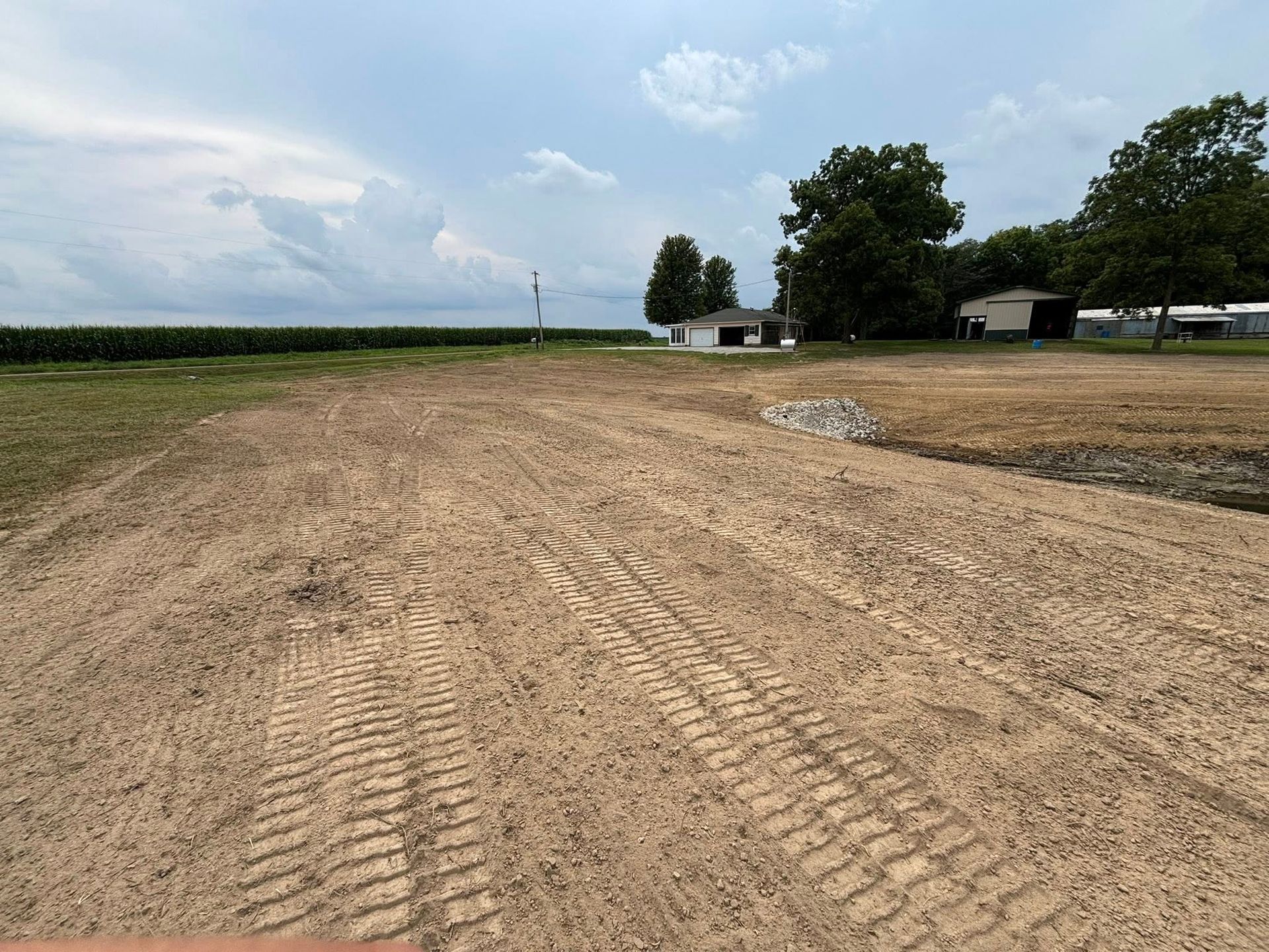 Dirt road with tire tracks leading towards a white building and green cornfield under a cloudy sky.