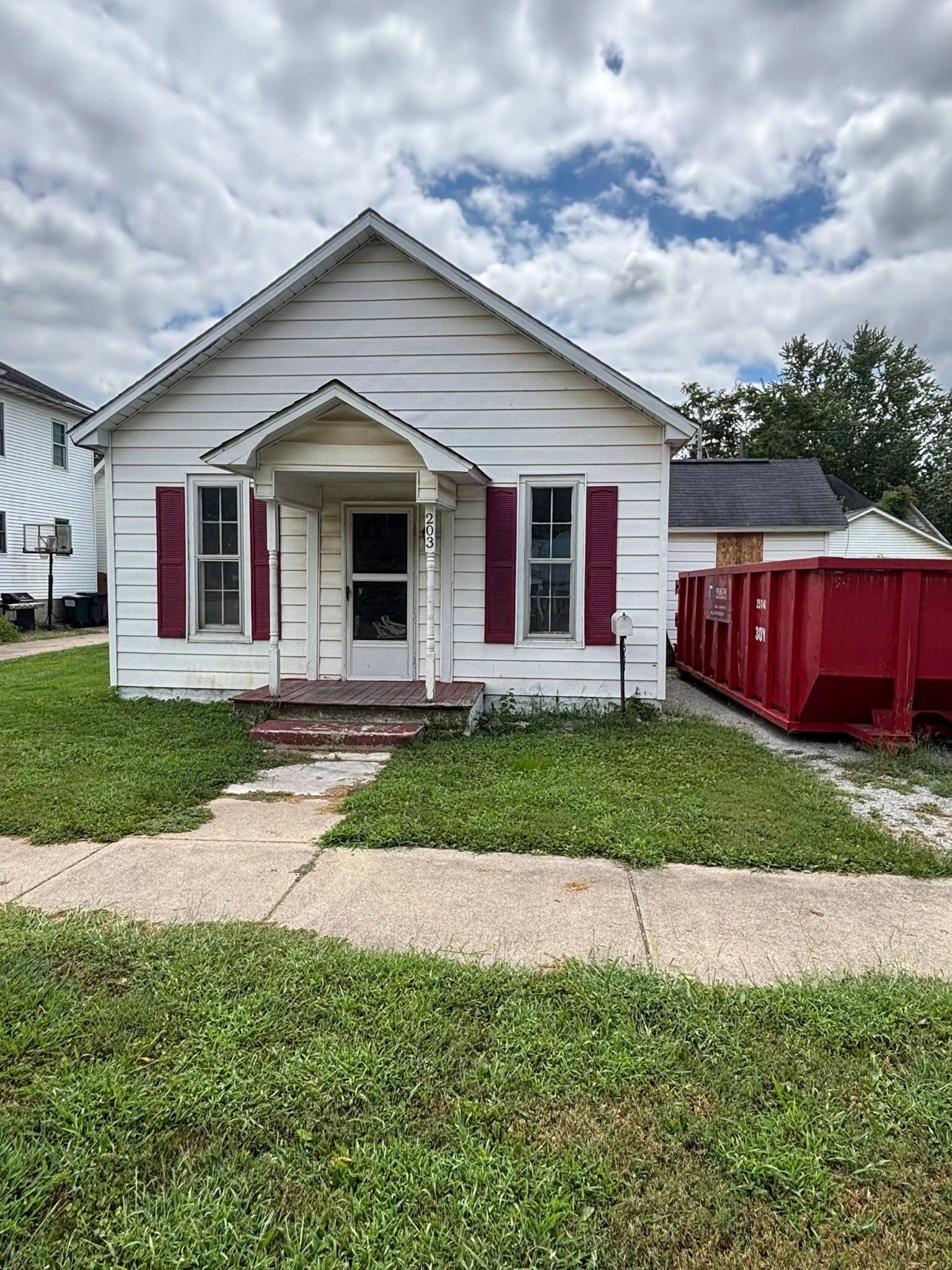 Small, white house with red shutters and a red dumpster next to it. Overgrown grass.