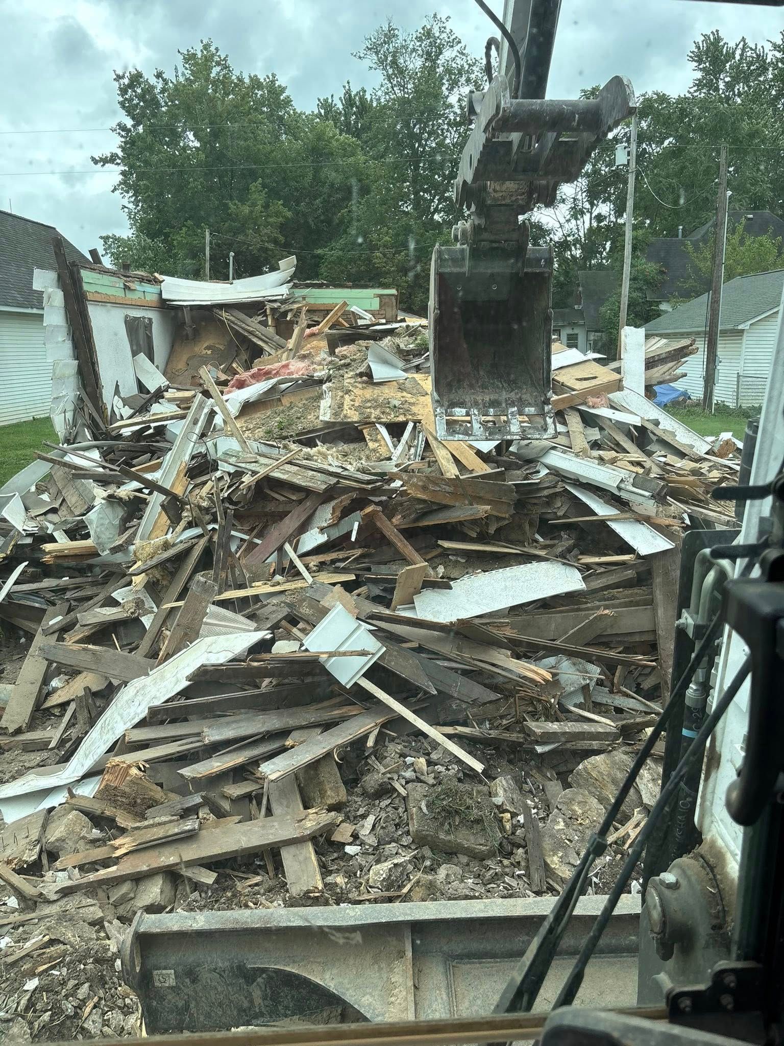 Demolition of a building. Excavator bucket scoops up debris of wood, walls, and roof. Exterior setting.