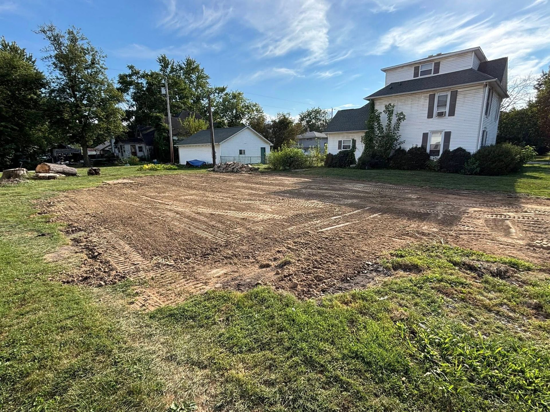 Cleared lot with dirt, grass, and a white house in the background under a blue sky.